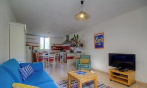 Ein Wohnzimmer mit einem blauen Sofa, einem Holztisch, einem Fernseher auf einem Schrank, einer Küche im Hintergrund und einem Essbereich mit Stühlen.