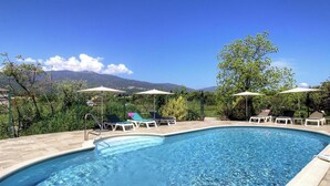Piscina con sillas de descanso y sombrillas, rodeada de vegetación y montañas al fondo.