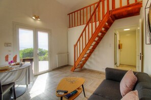 Ein Wohnzimmer mit einer Holztreppe, einer Essecke mit Tisch für zwei Personen und einem Sofa mit Kissen.
