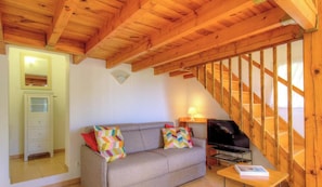 Ein Zimmer mit Holzbalkendecke, einer Couch mit bunten Kissen, einem Fernseher und einem kleinen Tisch mit einem Buch.