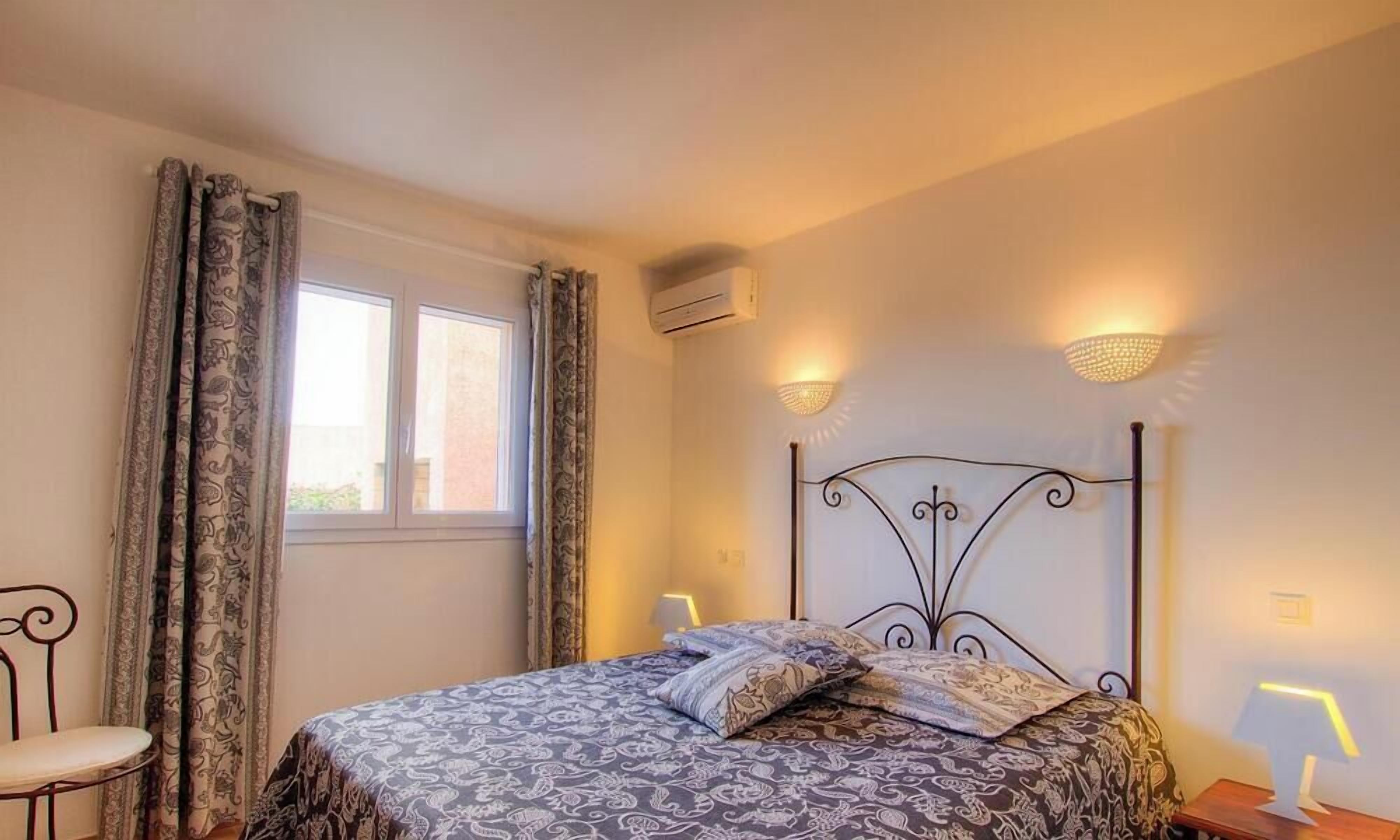 A bedroom with a patterned bedspread, a metal headboard, two wall-mounted lamps, a window with curtains, and an air conditioning unit.