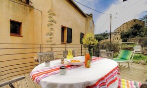 Ein runder Tisch mit gestreifter Tischdecke, eine Schüssel Orangen, eine Saftflasche und ein Krug auf einem Balkon mit Blick auf ein rustikales Gebäude und Grünflächen.