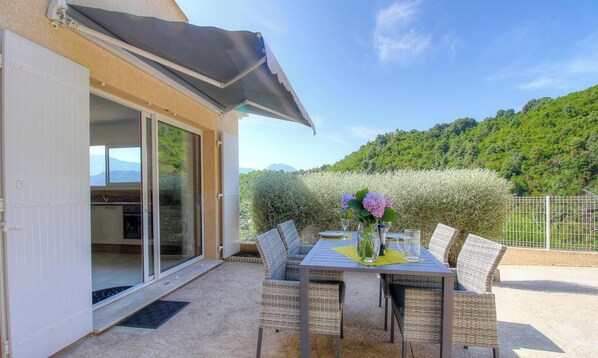 Un patio con una mesa lista para dos personas, una puerta corrediza de vidrio que da a una cocina y vistas a una ladera verde.