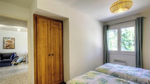 Ein Schlafzimmer mit einem hölzernen Kleiderschrank, einem Bett mit blattgemustertem Überwurf, einem Fenster mit Vorhängen und einer Pendelleuchte.