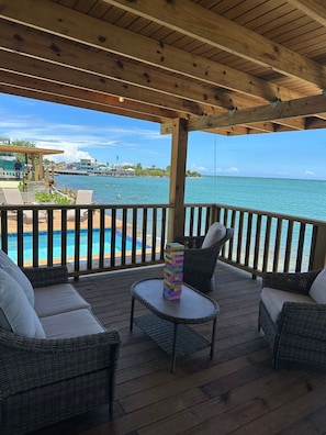 Eine Holzterrasse mit Korbmöbeln, mit Blick auf ein Schwimmbecken und das Meer.