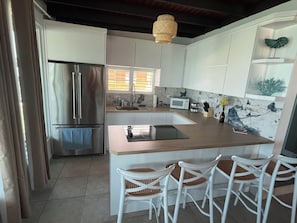 A modern kitchen with white cabinets, a stainless steel refrigerator, a microwave, and a sink. There are three white chairs with woven seats.