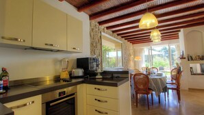 A kitchen with a stone wall, wooden ceiling beams, a dining area with a table and chairs, and a view of greenery outside. - The villa - AFA - Gîte (AFA)