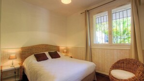 A bedroom with a bed, two bedside lamps, a wicker chair, a window with a view of greenery, and a ceiling light fixture. - The villa - AFA - Gîte (AFA)