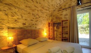 Ein Schlafzimmer mit Steinwand, Holz-Kopfteil, Nachttischlampen, einem Bett mit Kissen, einem hölzernen Regal und einem Fenster mit Vorhängen.