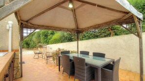 Een terras buiten met een overdekte zithoek, een tafel en stoelen.