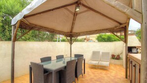 Eine überdachte Terrasse mit Glastisch, Korbstühlen, Grillbereich und Blick auf die umliegende Grünfläche.