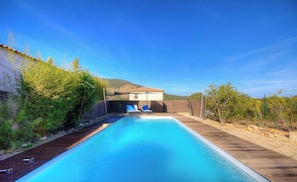 Una piscina rettangolare con un solarium in legno, un'area relax coperta e montagne sullo sfondo.