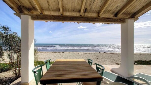 Ein Holztisch mit Stühlen unter einer Holzpergola mit Blick auf den Strand und das Meer.