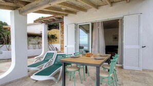 Eine Außenpatio mit Tisch und Stühlen, ein überdachter Bereich mit Holzbalkendecke und eine Glasschiebetür, die in einen Innenraum führt.