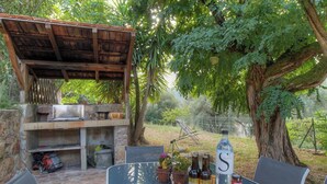 Een terras buiten met een steen barbecue, zitplaatsen en een grote boom.