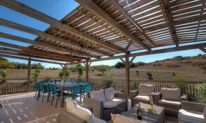 Eine Terrasse im Freien mit einer Holz-Pergola, grünen Stühlen, Korbsesseln und einem Essplatz.