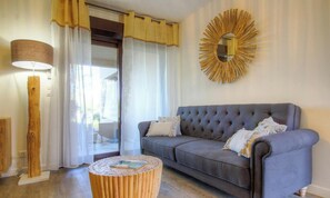 Ein Wohnzimmer mit grauem Sofa, einem runden Holztisch, einer Stehlampe und einem Spiegel in Sonnenform an der Wand.