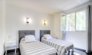 Ein Zimmer mit zwei Betten, jeweils mit grauem Kopfteil und weißer Bettwäsche, einem kleinen Nachttisch mit Lampe und einem Fenster mit Vorhängen.