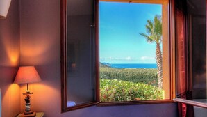 Ein Zimmer mit Fenster und Blick auf eine Landschaft mit Palme und Meer.