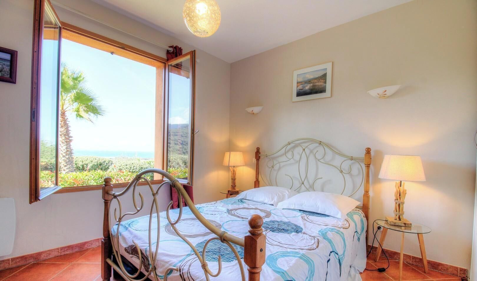 A bedroom with a bed, two bedside lamps, a painting on the wall, and a window with a view of greenery.