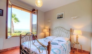 A bedroom with a bed, two bedside lamps, a painting on the wall, and a window with a view of greenery. - Le 693 - PROPRIANO - Gîte (PROPRIANO)