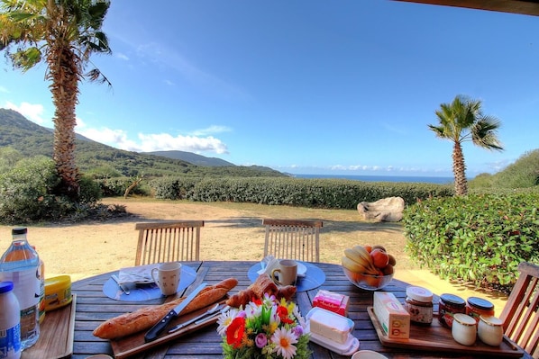 A table set outdoors with food, a bottle, and a view of the sea and hills. - Le 693 - PROPRIANO - Gîte (PROPRIANO)