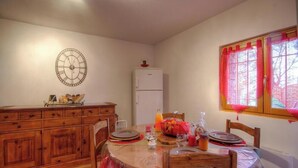 Uma sala de jantar com uma mesa de madeira, cadeiras, um relógio na parede, um refrigerador e uma janela com cortinas.
