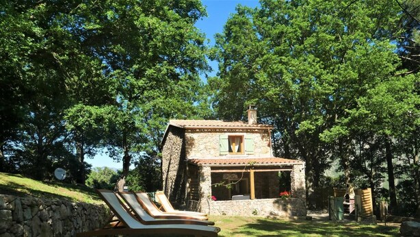 Uma casa de pedra com telhado de telhas, rodeada por árvores. Existem duas cadeiras de descanso em primeiro plano.