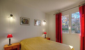 Ein Schlafzimmer mit einem Bett, zwei Nachttischlampen, zwei gerahmten Bildern, einem Fenster mit roten Vorhängen und Blick auf Bäume.