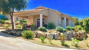 Ein Bungalow mit überdachter Veranda, einer Steinmauern und verschiedenen Pflanzen und Sträuchern.