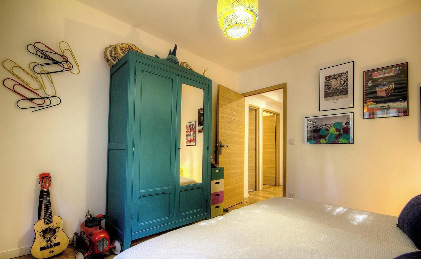 A bedroom with a bed, a guitar, a teal wardrobe, and a door leading to another room.