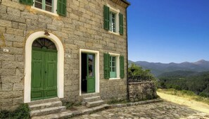 Un edificio de piedra con contraventanas verdes y una vista despejada de montañas al fondo.