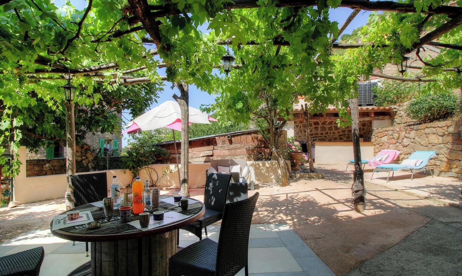 Une terrasse extérieure avec une table ronde et des chaises, entourée de verdure et d’un mur en pierre.