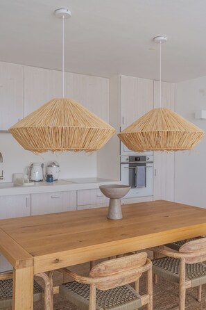 Une table à manger en bois, des chaises à assise tressée, un grand saladier au centre et deux suspensions décoratives tressées.