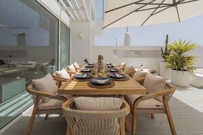 A sunlit outdoor dining area with a wooden table, cushioned chairs, and a large umbrella.