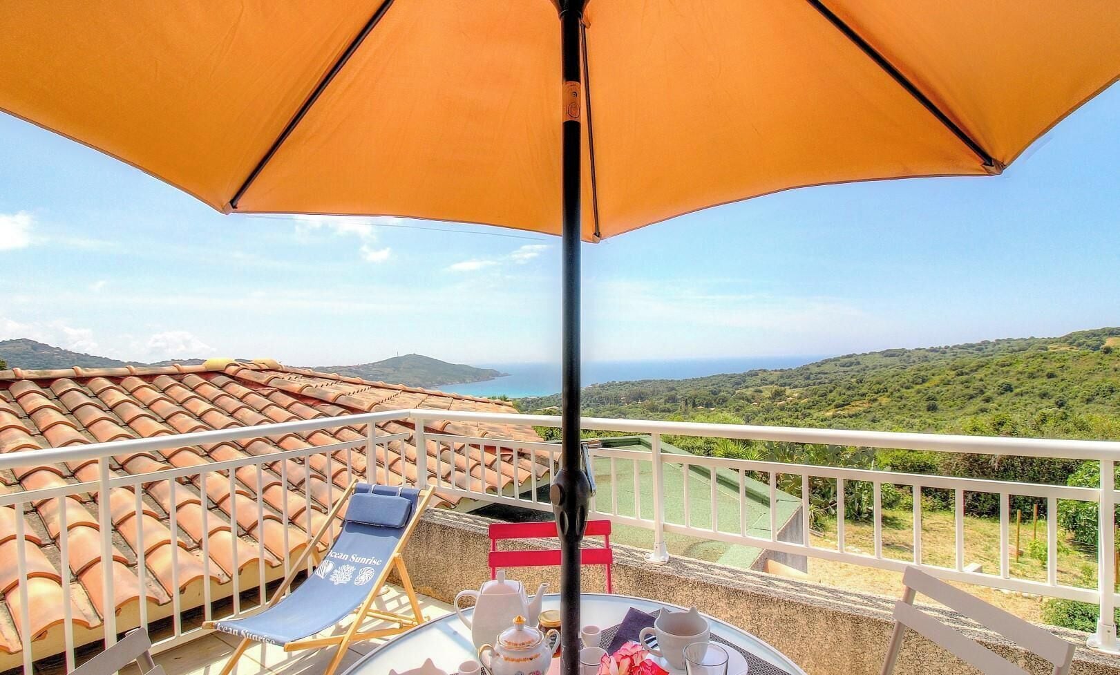 A balcony with a table set for two, a sun umbrella, and a view of a green landscape.