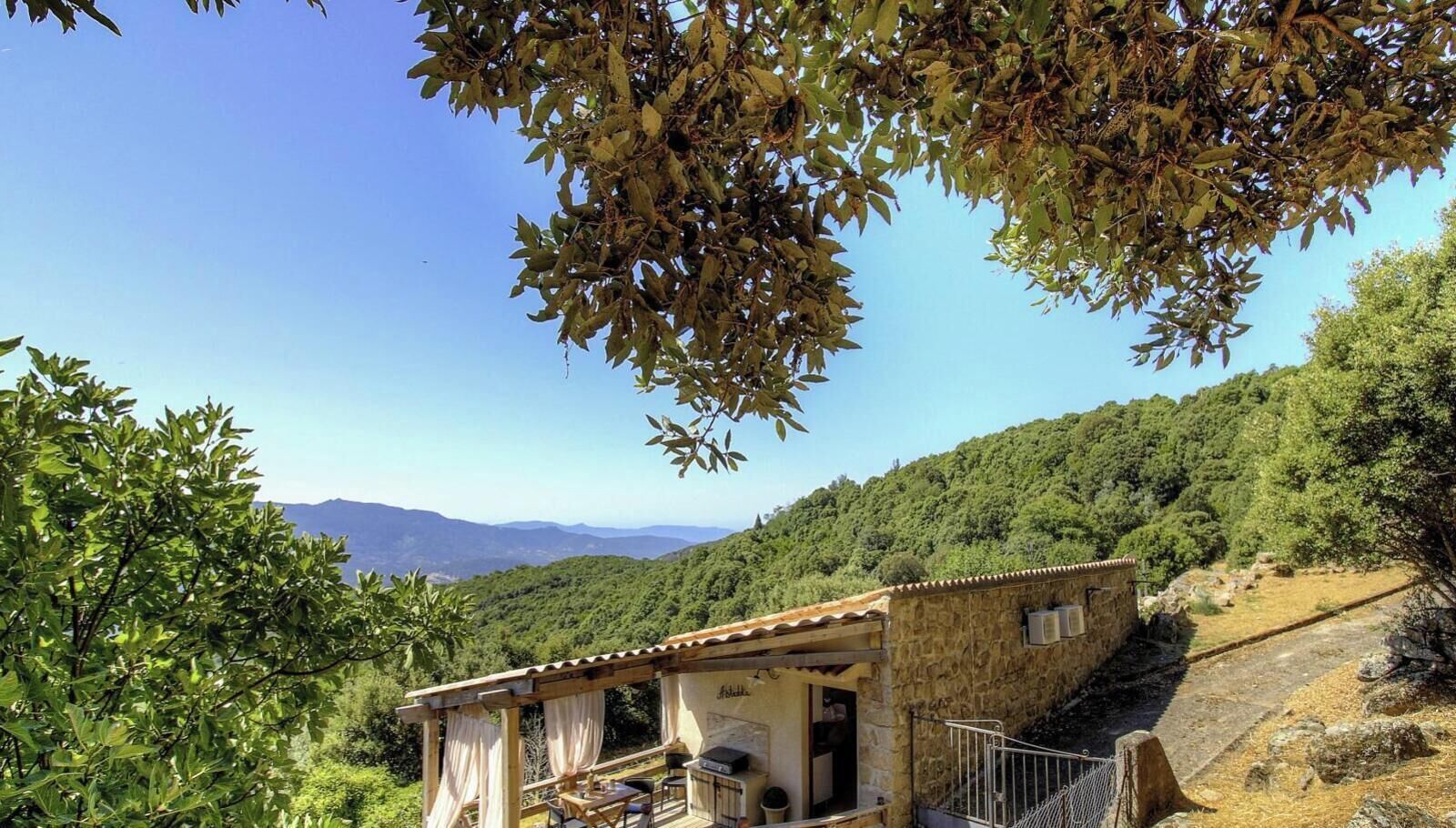 Un edificio de piedra con balcón, rodeado de vegetación y montañas al fondo.