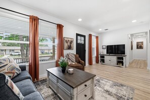 A modern living room with a sofa, coffee table, and TV stand, large windows with curtains, and a door.