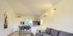 A modern kitchen with a central island, bar stools, and a grey sofa in a bright living area. - Villa Léa - SAINTE LUCIE DE PORTO VECCHIO (SAINTE LUCIE DE PORTO VECCHIO)