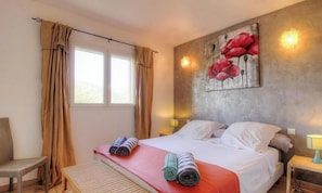A hotel room with a bed, two bedside lamps, a window with curtains, a chair, and a wall with a floral painting. - Villa Léa - SAINTE LUCIE DE PORTO VECCHIO (SAINTE LUCIE DE PORTO VECCHIO)