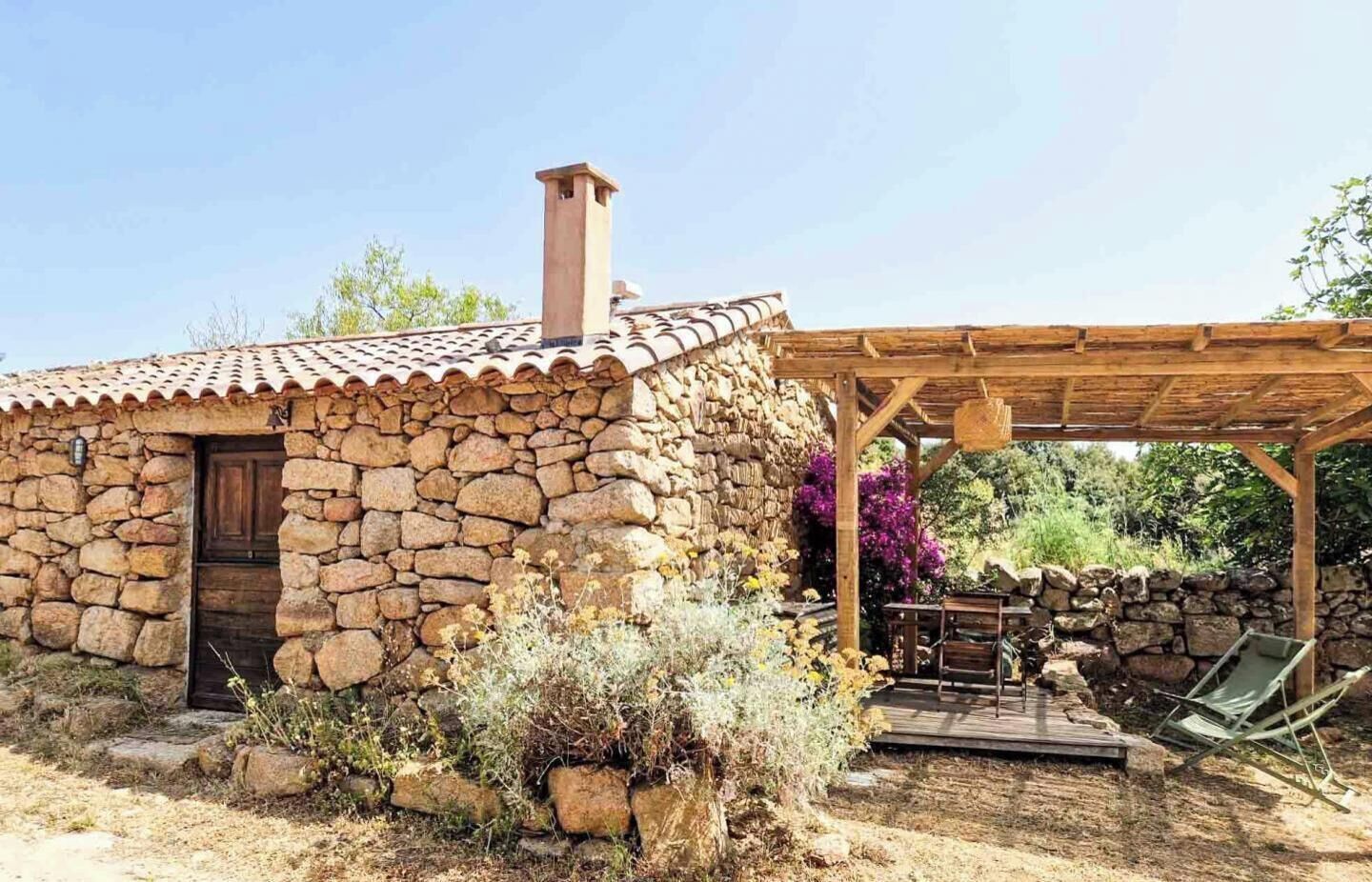 Una casa in pietra con una porta in legno, un camino e una zona salotto esterna coperta.