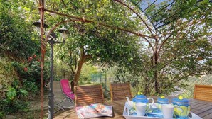 Een houten terras met een theetafel, omringd door stoelen, onder een pergola begroeid met klimplanten.