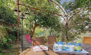 Eine Holzterrasse mit einem für Tee gedecktem Tisch, umgeben von Stühlen, unter einer mit Kletterpflanzen bewachsenen Pergola.
