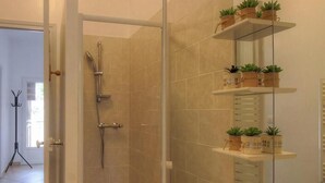 A bathroom with a glass-enclosed shower, a sink with a mirror, and shelves with potted plants. - Villa Charlain - PIETROSELLA (PIETROSELLA)
