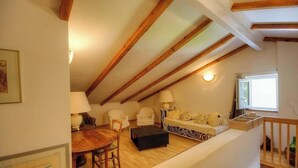 Een loft-achtige kamer met zichtbare houten balken, een bank, stoelen, een tafel en een raam.