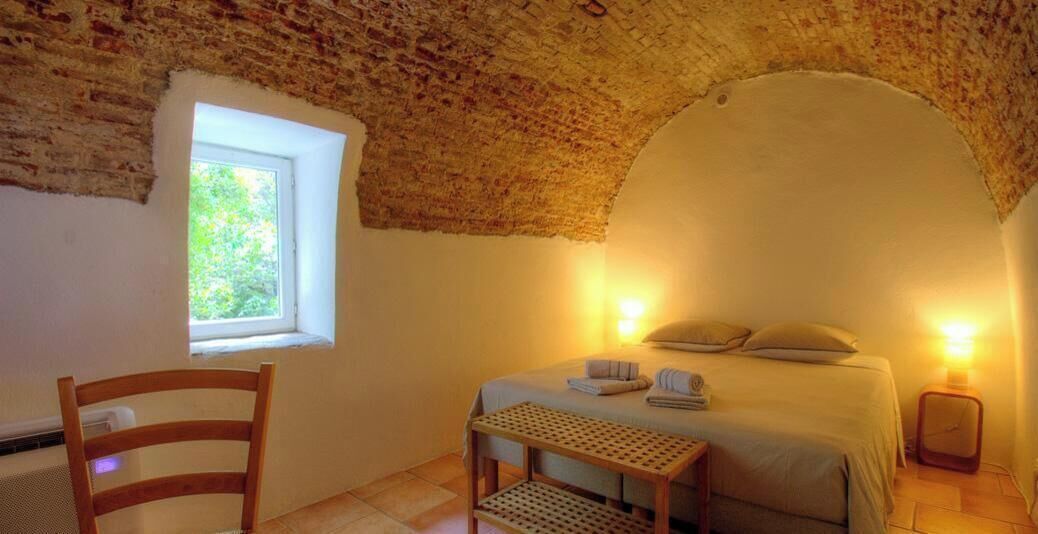 A room with a bed, a chair, a small table, a window, and a brick ceiling.