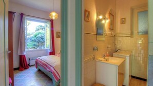 Un dormitori amb un llit, una finestra amb cortines, una làmpada penjant i un bany amb un lavabo, mirall i barra de tovalloles.
