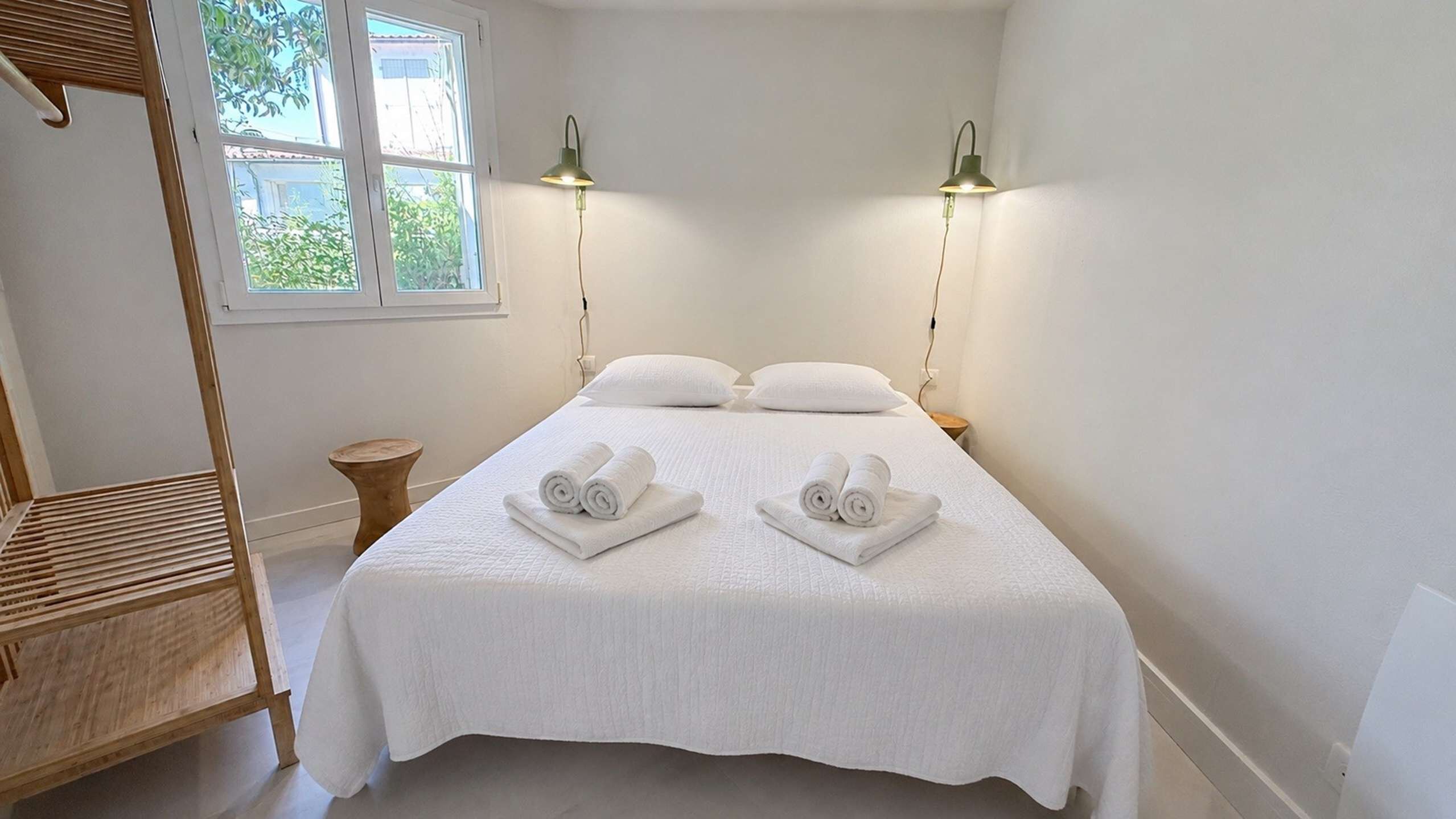 Een net opgemaakt bed met opgerolde handdoeken, twee bedlampen, een houten bankje en een raam met uitzicht op groen.
