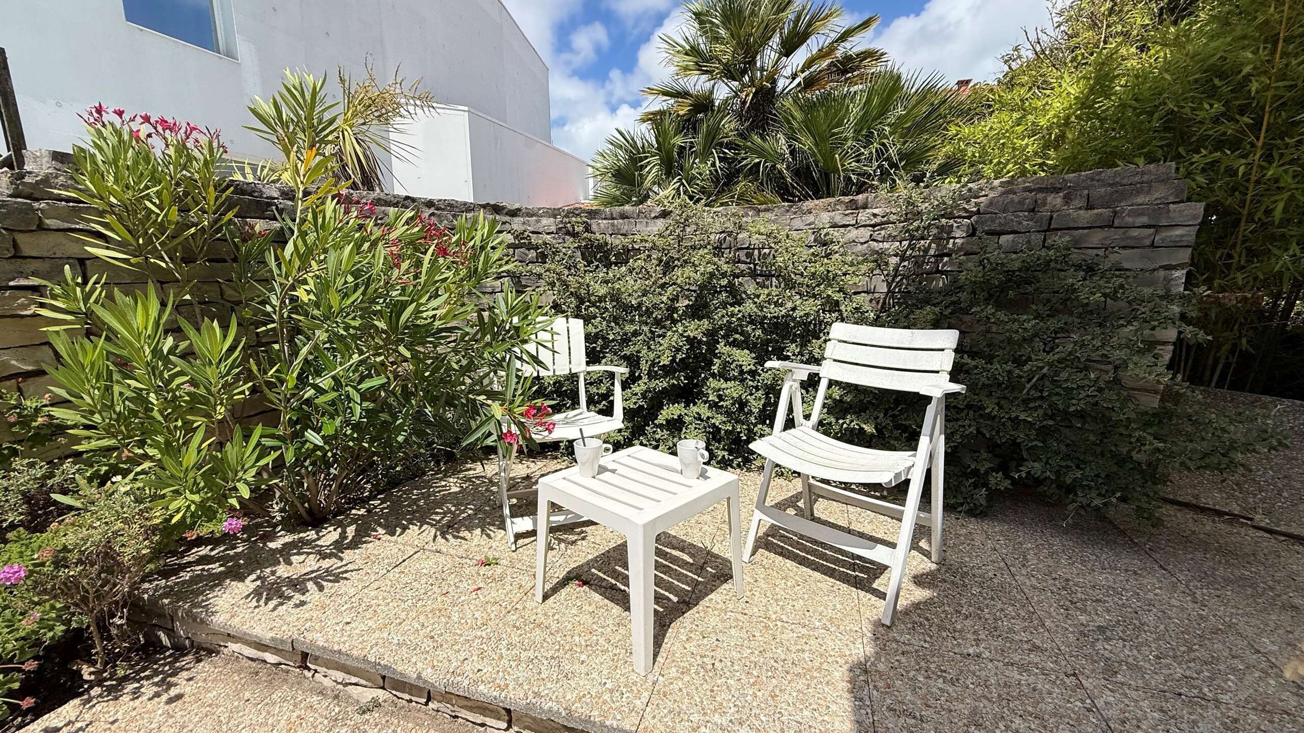 A patio with two white chairs and a table, surrounded by greenery and a stone wall.