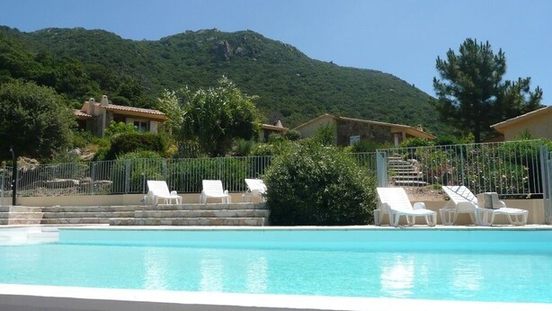 Una piscina con sillas de descanso blancas, una escalera de piedra y edificios al fondo.
